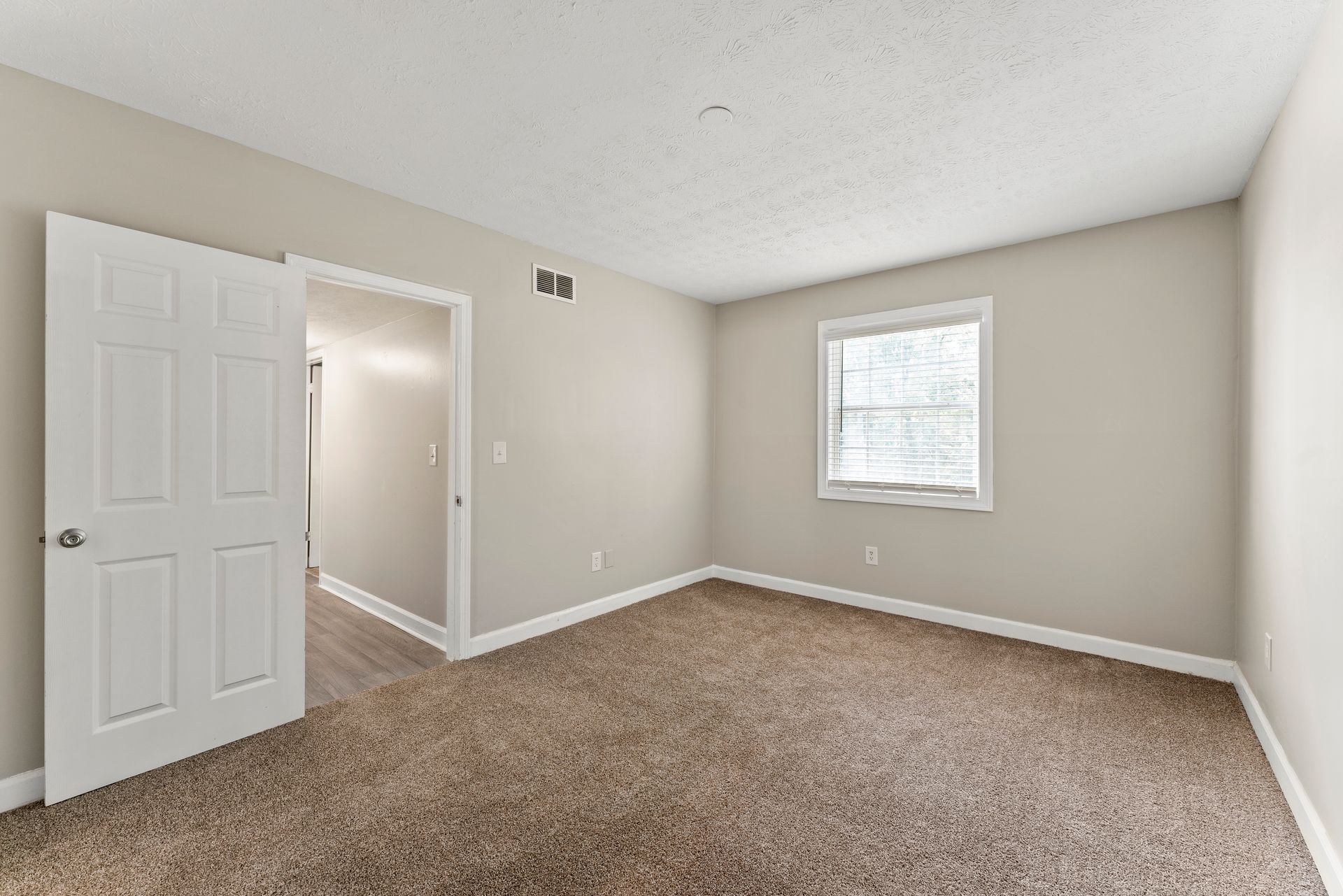 Empty beige bedroom with carpet, open white door, and a small window with blinds.