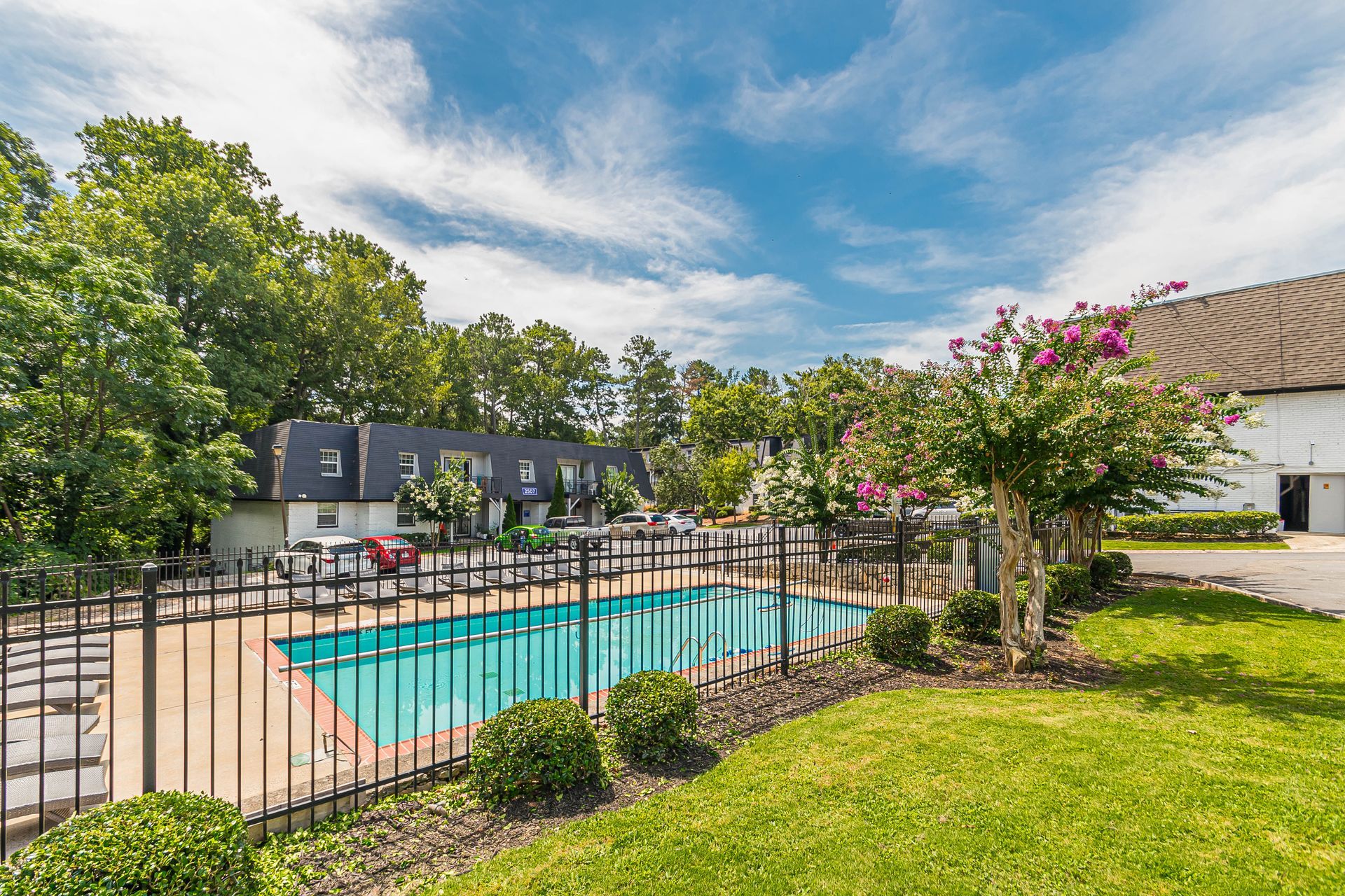 Outdoor apartment pool with turquoise water, black fence, green lawn, and flowering trees under a blue sky