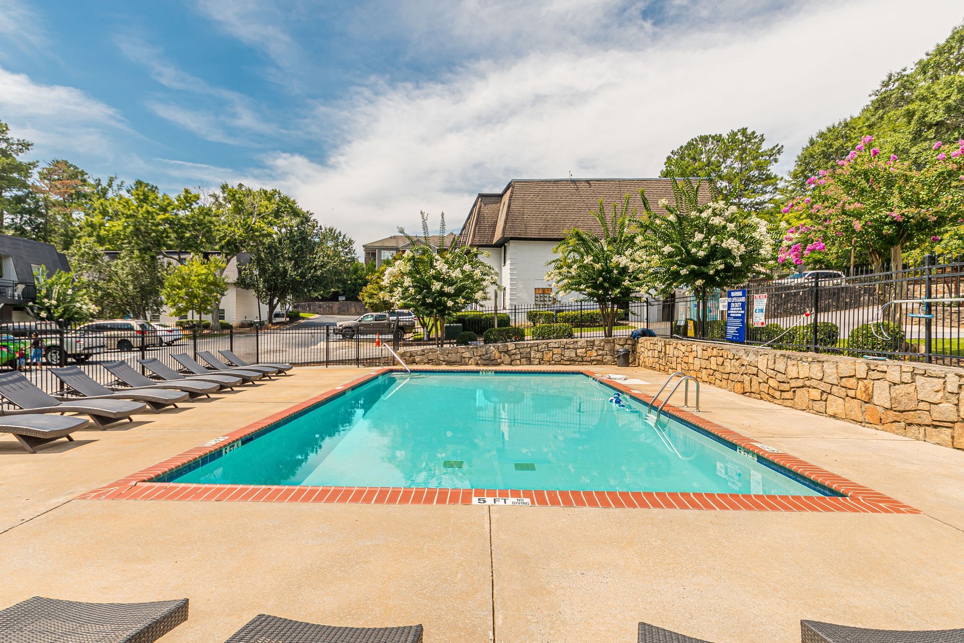 Outdoor swimming pool with lounge chairs, stone deck, and trees under a partly cloudy sky