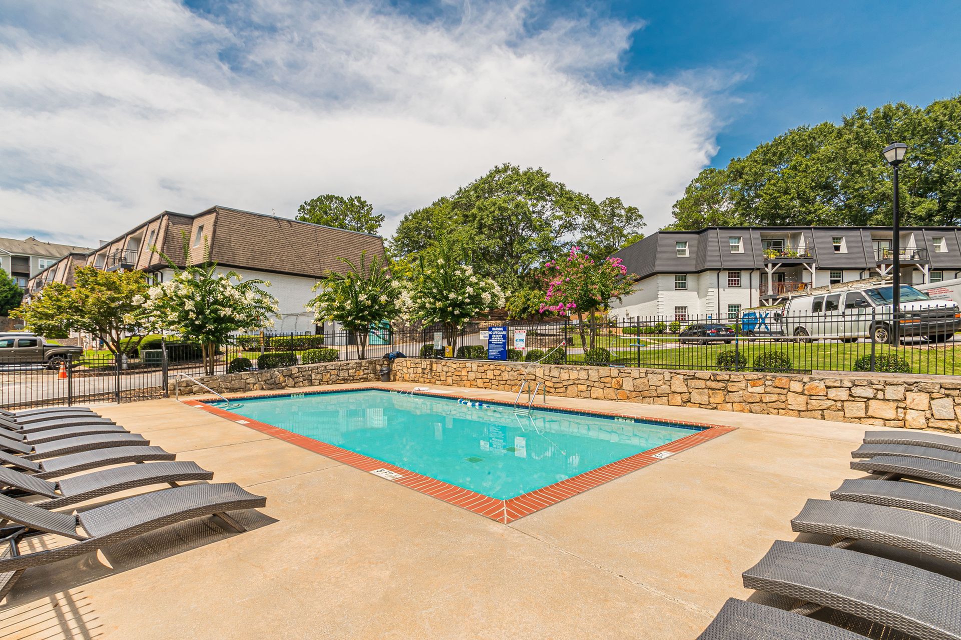 Outdoor swimming pool with lounge chairs, stone wall, and apartment buildings under a partly cloudy sky