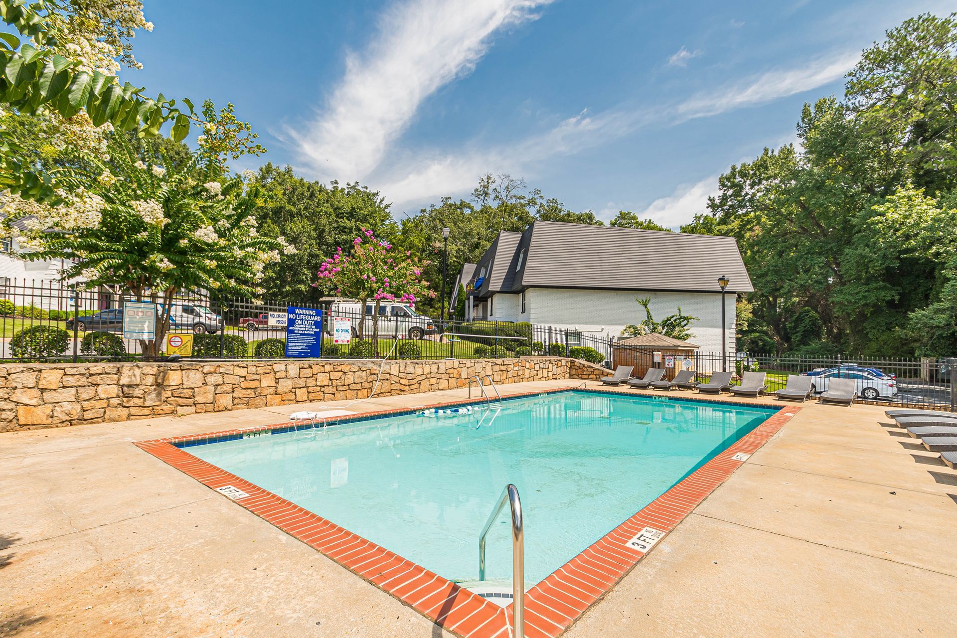 Outdoor swimming pool with turquoise water, tan deck, stone wall, and trees under a blue sky