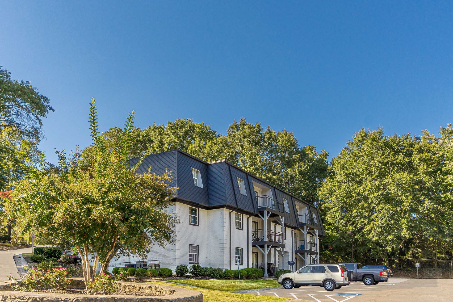 Apartment building with dark roof, parked cars, and trees under a clear blue sky