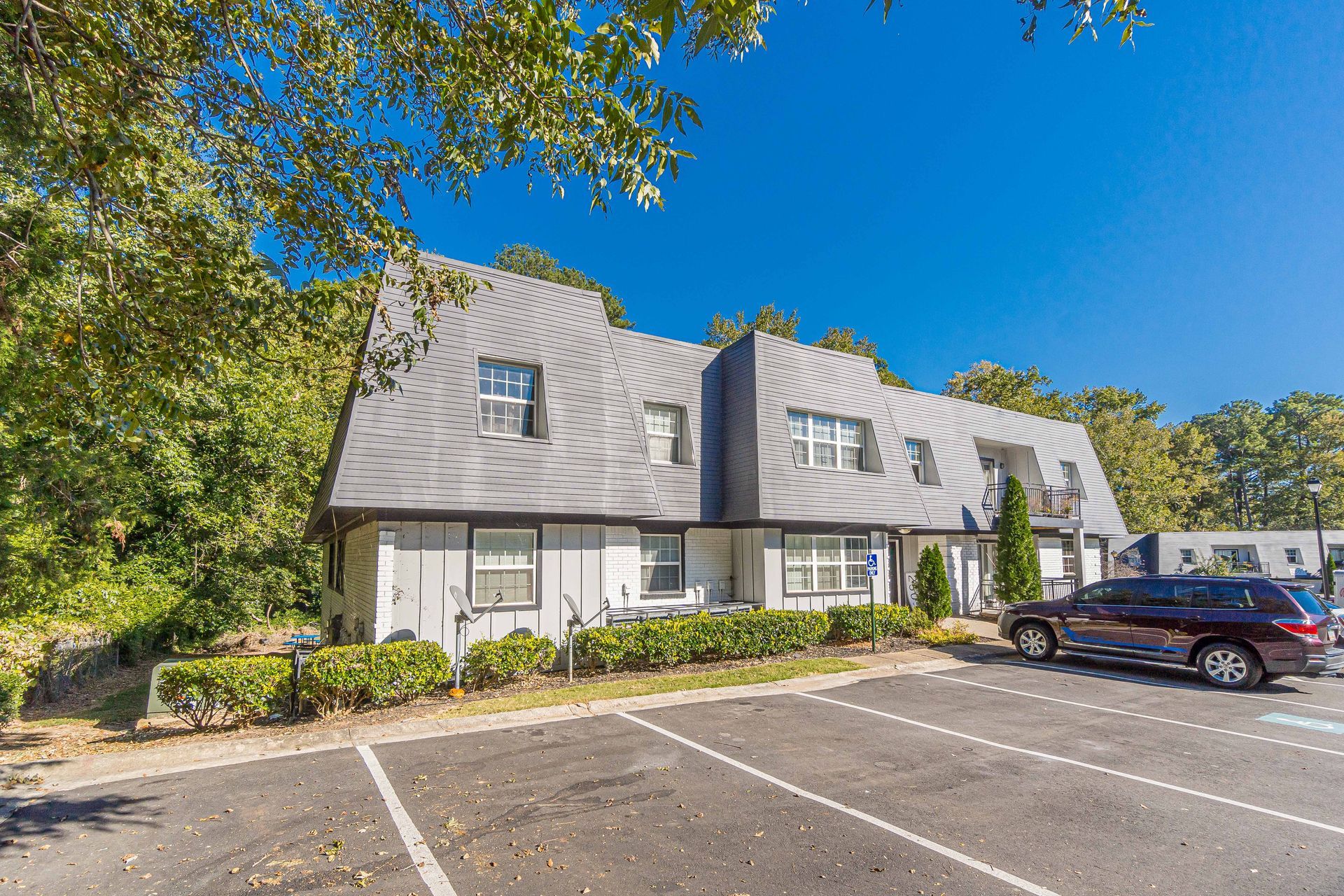 Apartment building with gray roof, trees, and parked cars in a lot under a blue sky