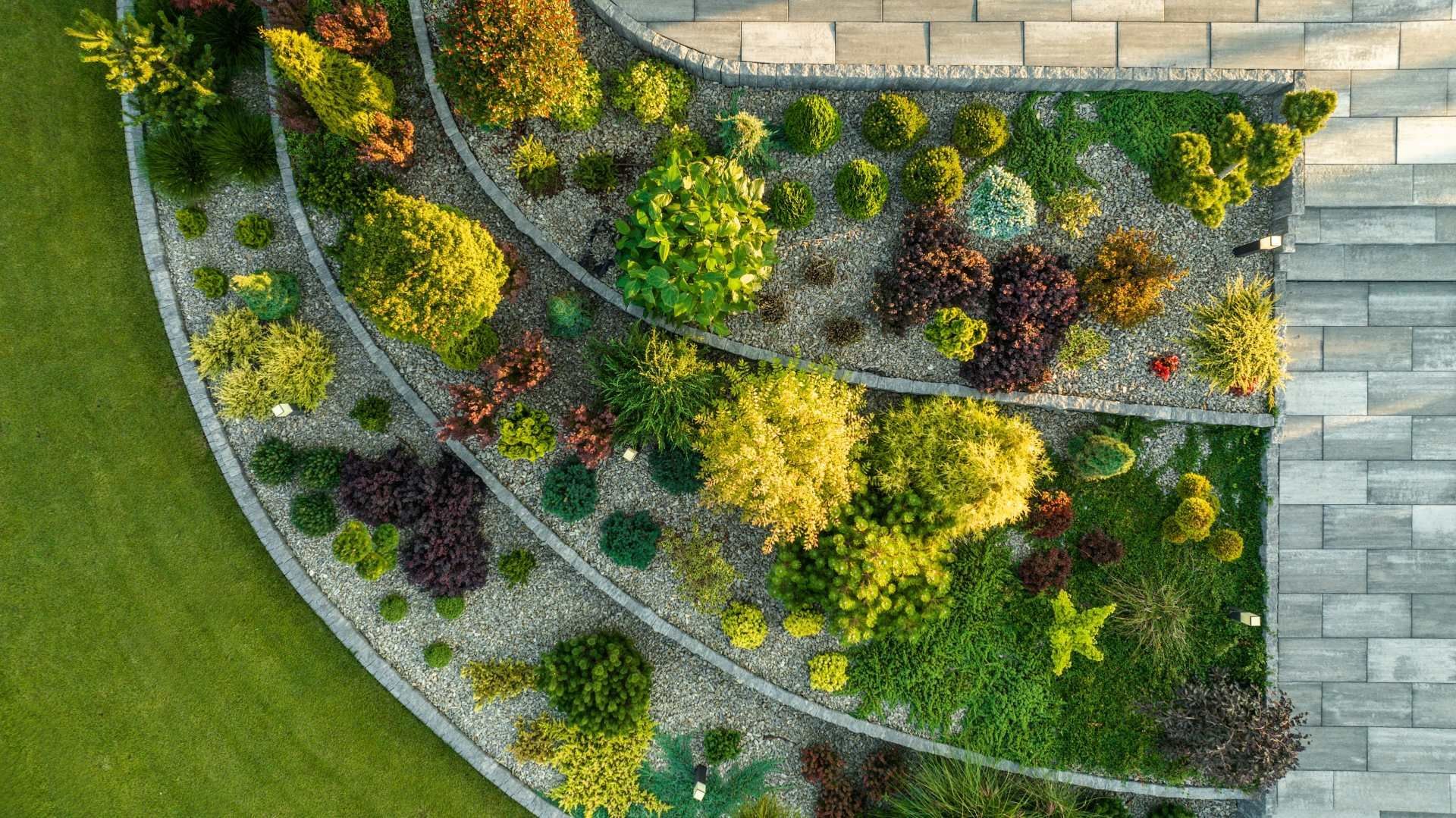 Aerial view of a curved landscaped garden bed with colorful shrubs beside a stone patio and lawn