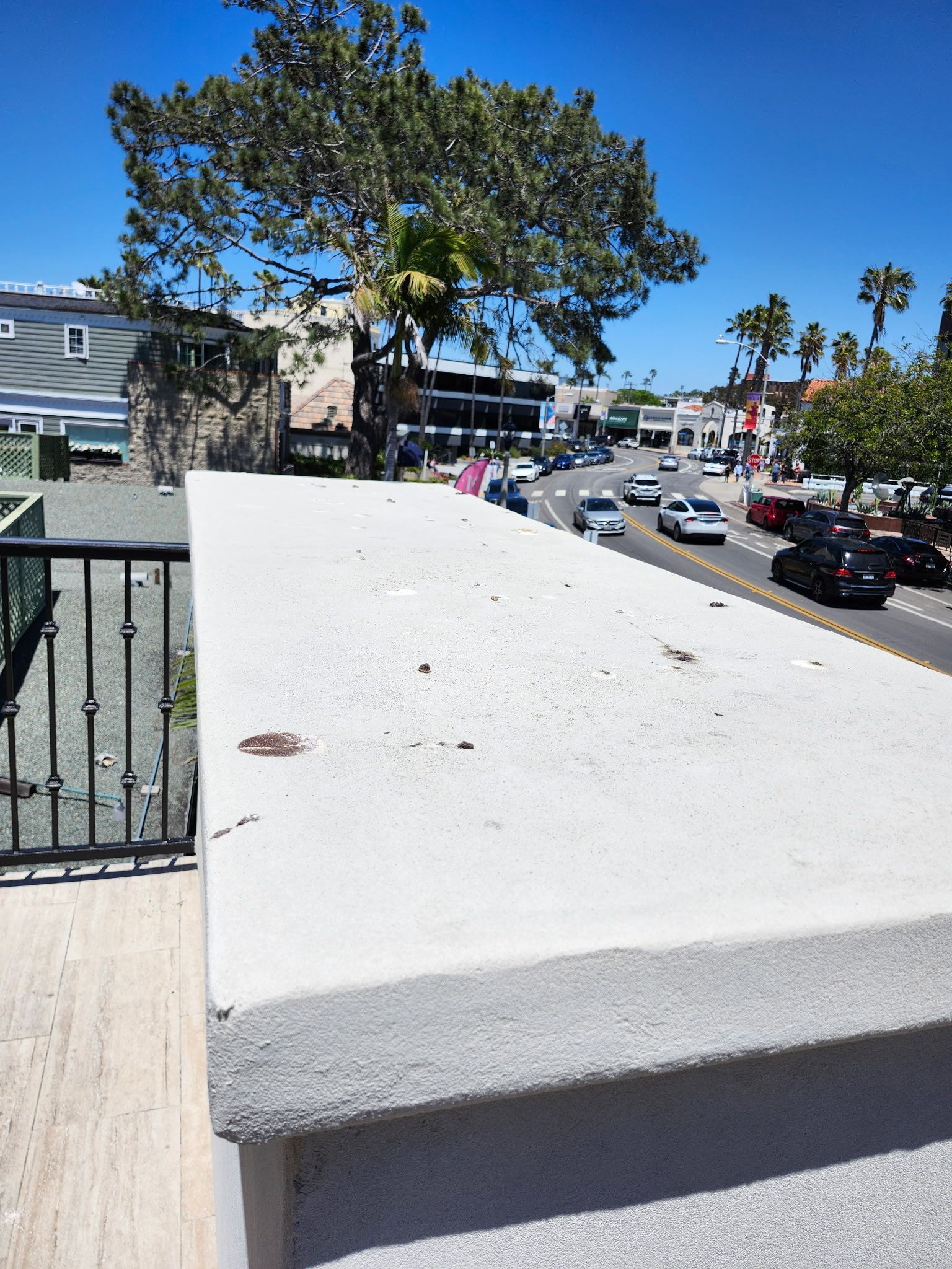 Sunny street view from a rooftop terrace with white railing, trees, parked cars, and houses below.