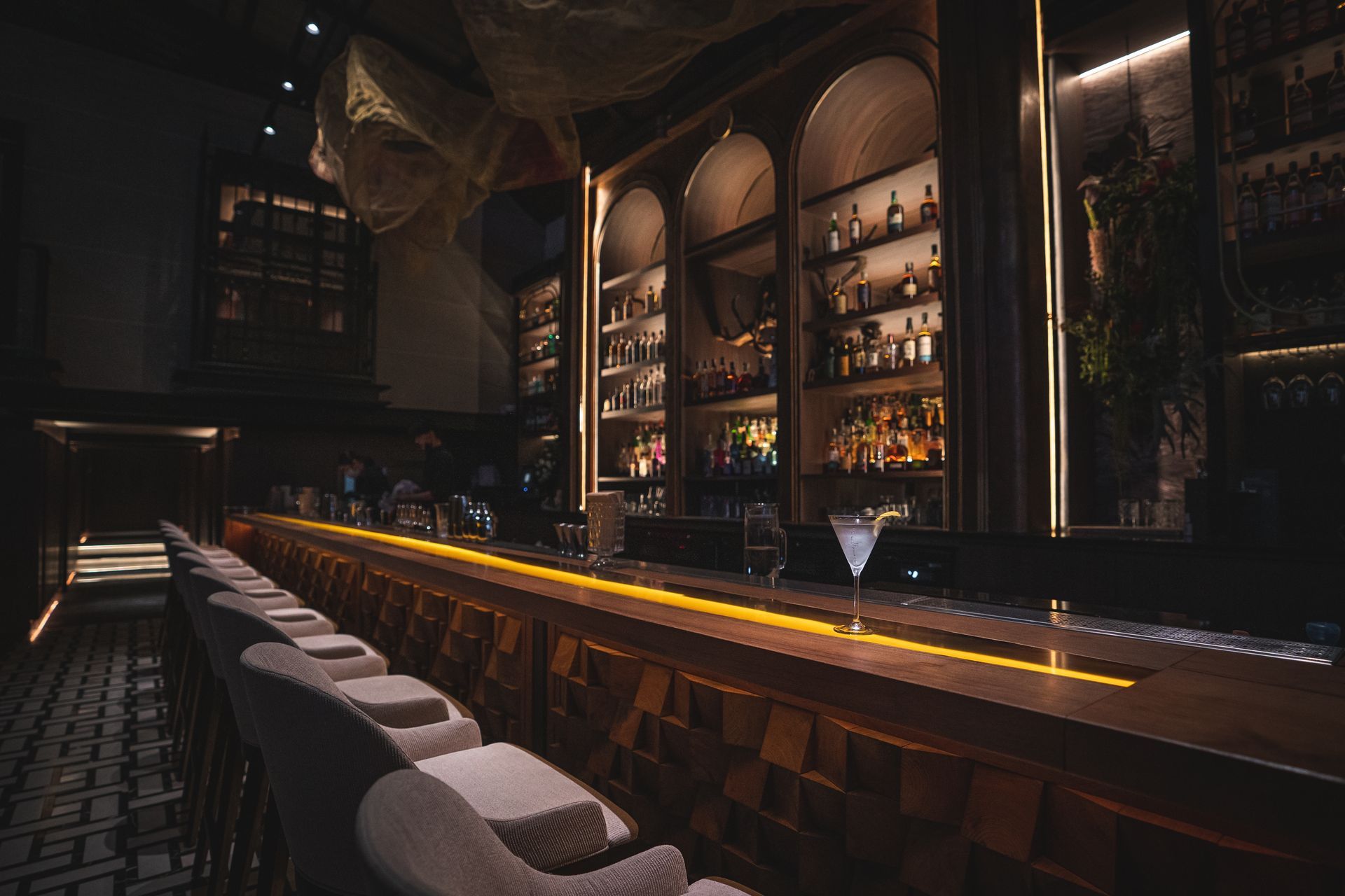 Dimly lit upscale bar interior with a long counter, stools, and shelves of bottles