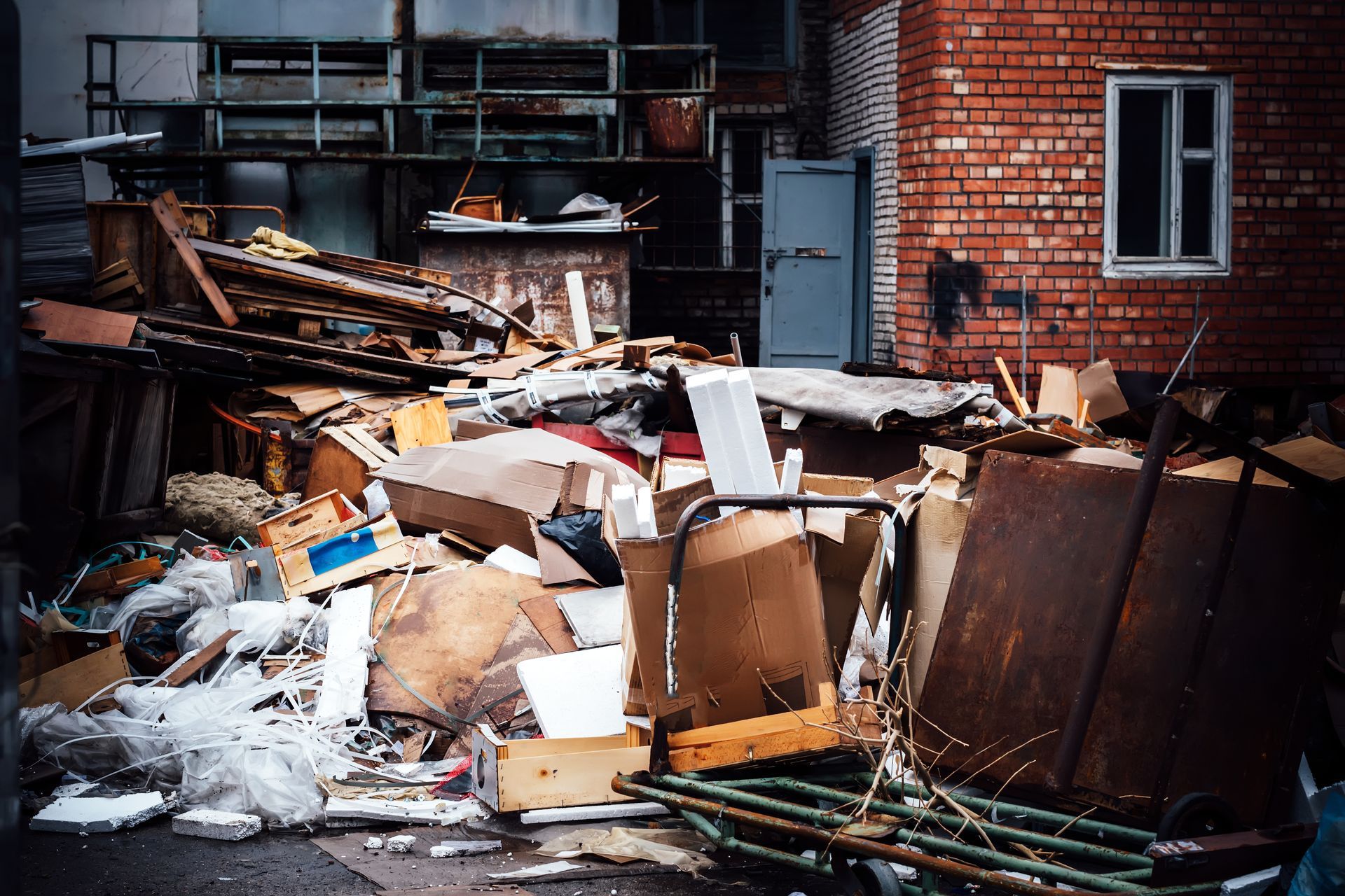 Cluttered alley with piles of broken furniture, cardboard, and trash beside a brick building