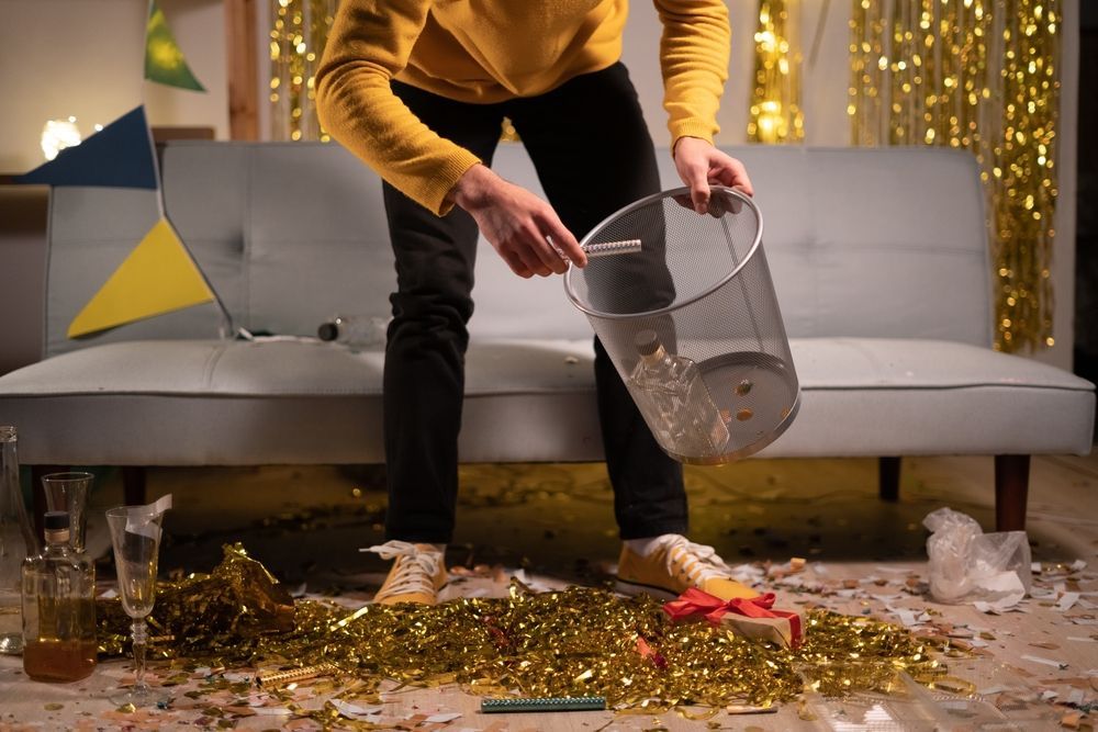 A man is throwing trash into a trash can in a messy living room.