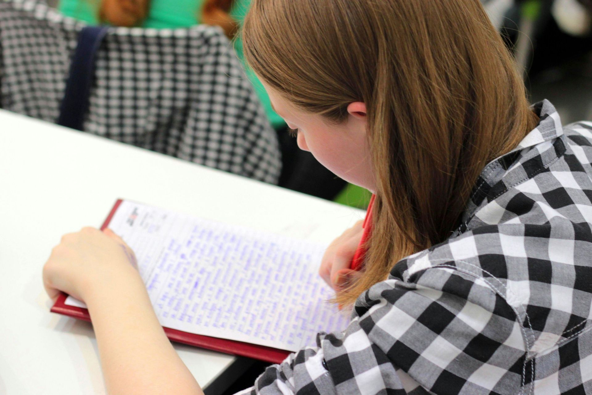 Woman with light brown hair writing on paper with a red pen, wearing a black and white checkered shirt.
