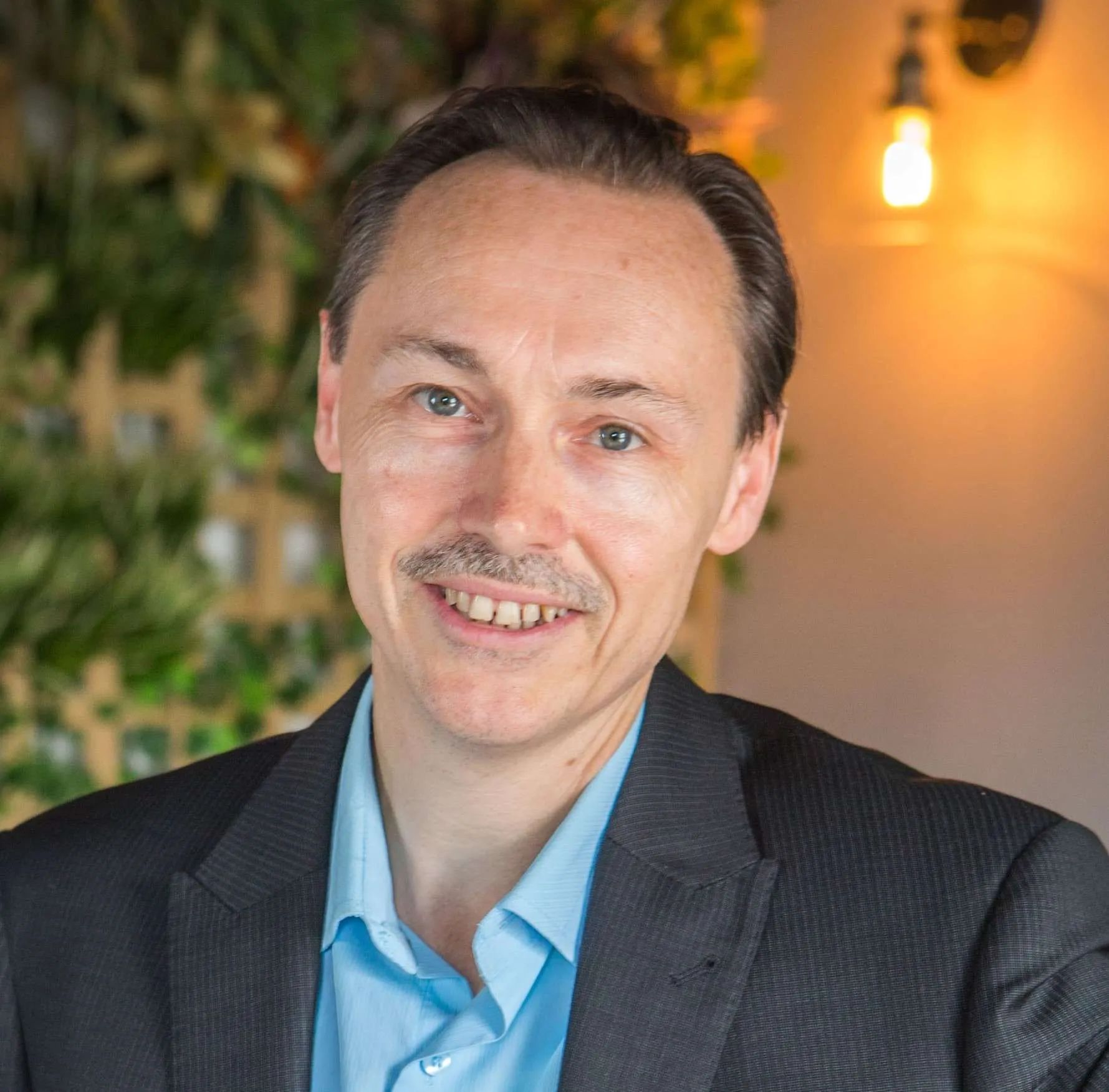 Smiling man in a dark blazer and light blue shirt, seated indoors with warm lights and greenery behind him