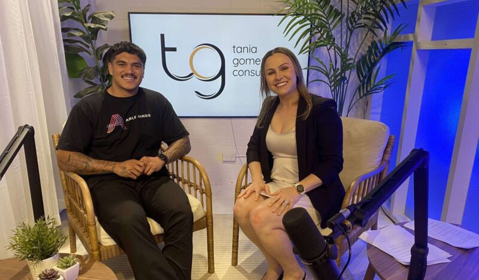 Two people seated in a studio, smiling beside a microphone and a screen with a “tg” logo.