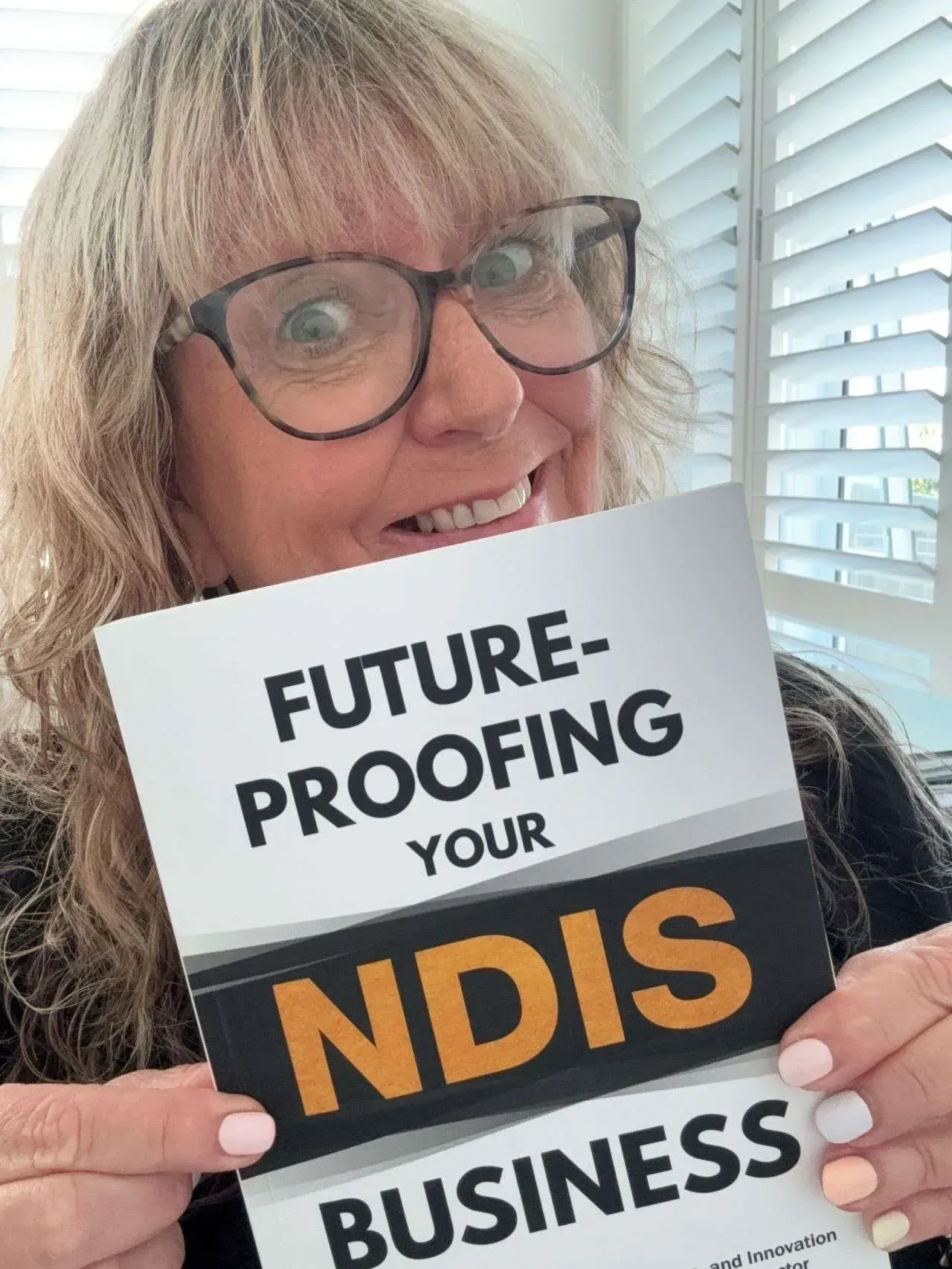 Smiling woman holding a sign reading “Future-proofing your NDIS business” indoors by a window.
