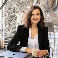 Woman in black blazer smiling at a café table with a laptop and coffee cup