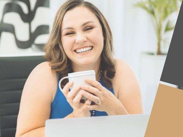 Smiling person holding a white mug at a laptop in a bright office setting