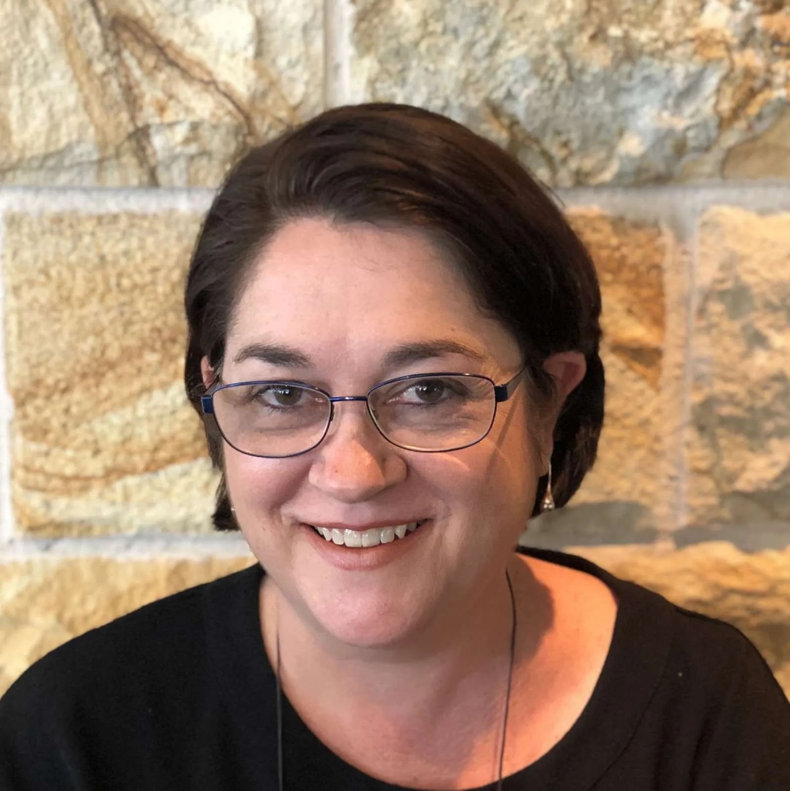 Smiling person with glasses in front of a stone wall