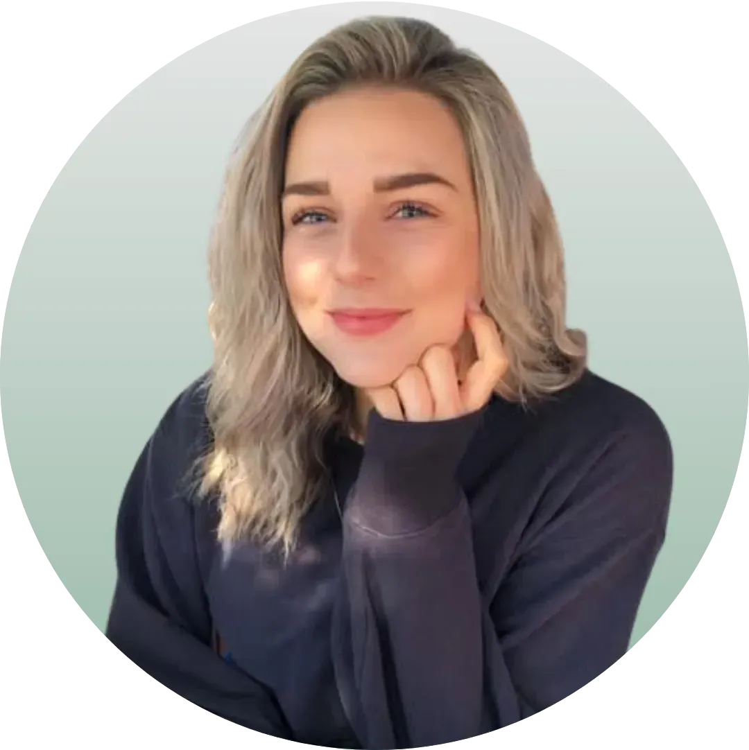 Portrait of a smiling blonde person in a dark sweater, resting their chin on their hand against a light background