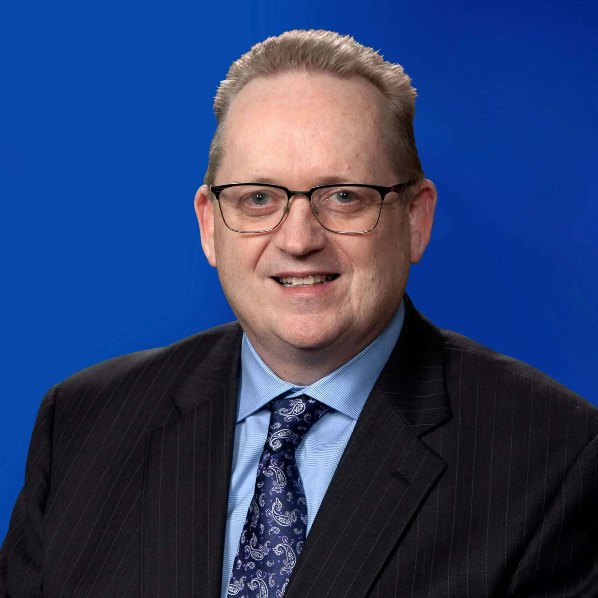 Studio headshot of a person in a dark suit and blue tie against a blue background