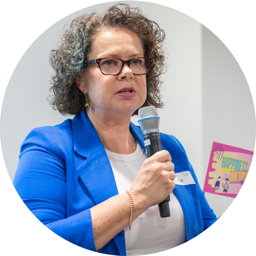 Woman in a blue blazer speaking into a microphone at an event, holding a small colorful booklet