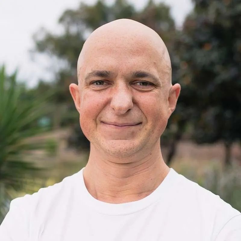 Smiling bald person in a white shirt outdoors with blurred greenery in the background