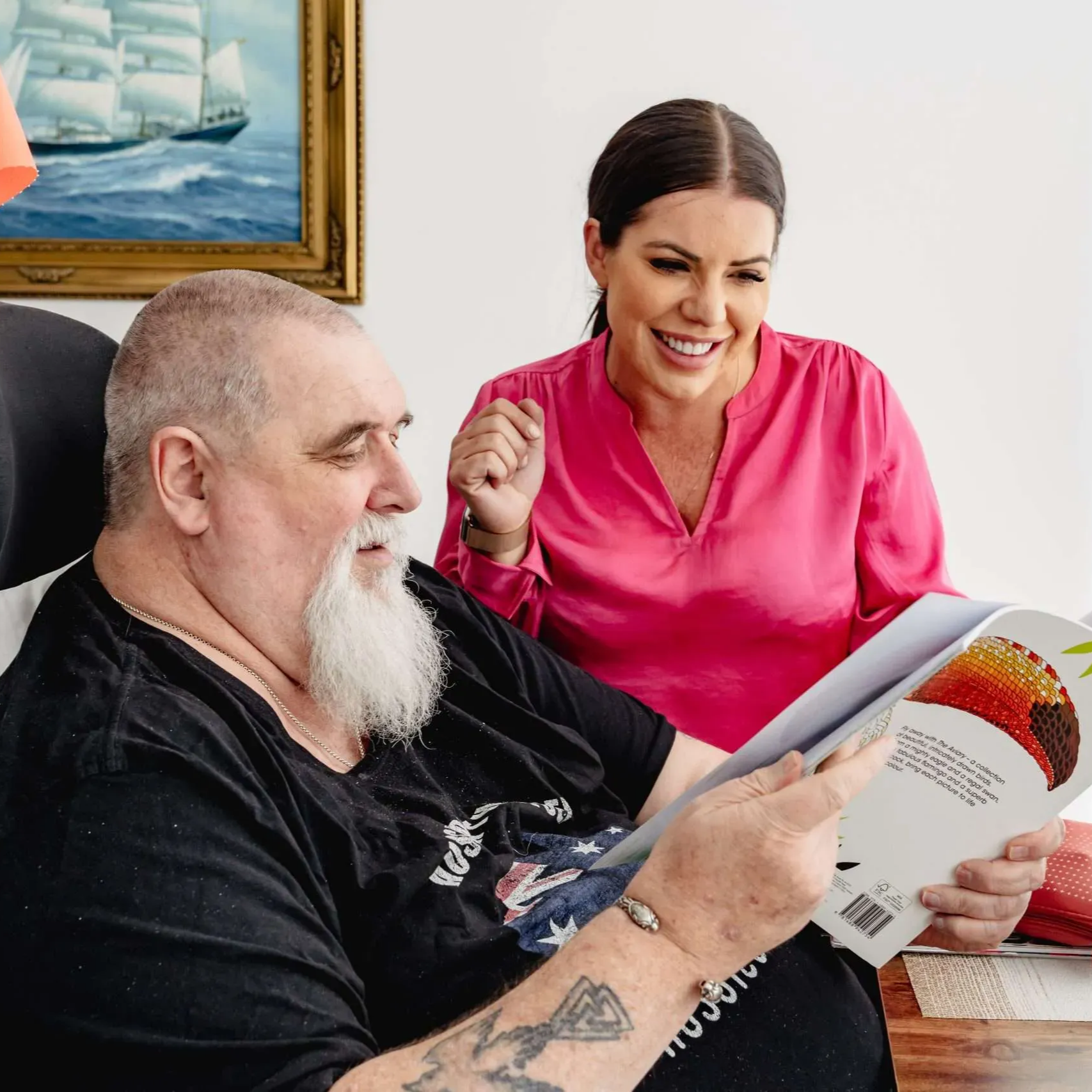 Man in black shirt reads a booklet while a smiling woman in pink stands beside him in a bright room.