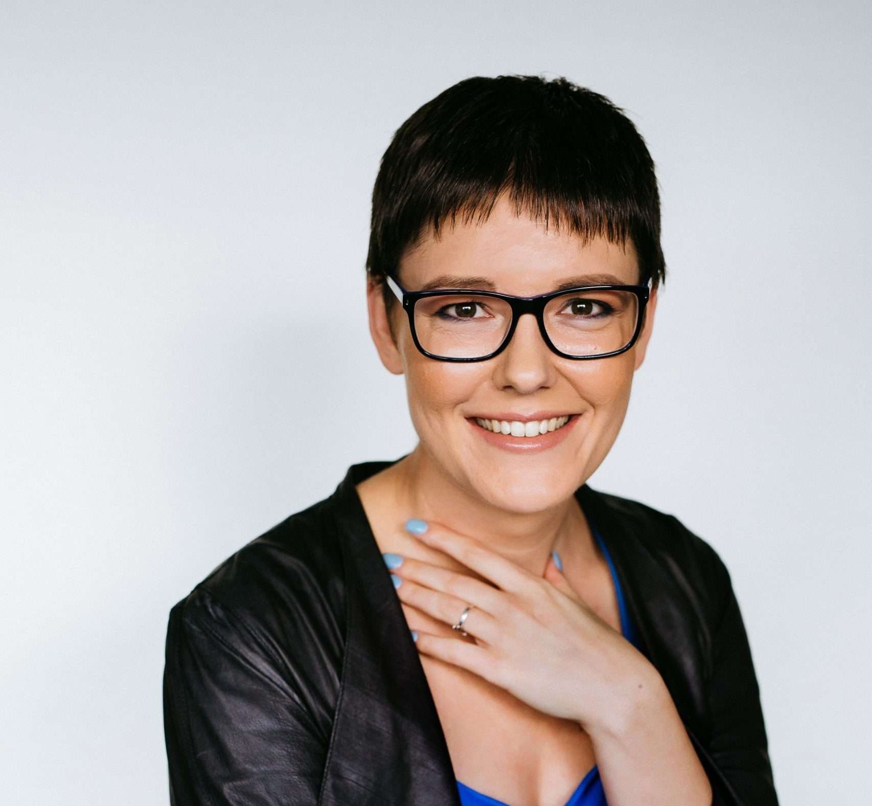 Smiling person with short dark hair and glasses, hand on chest, wearing a black jacket against a light background