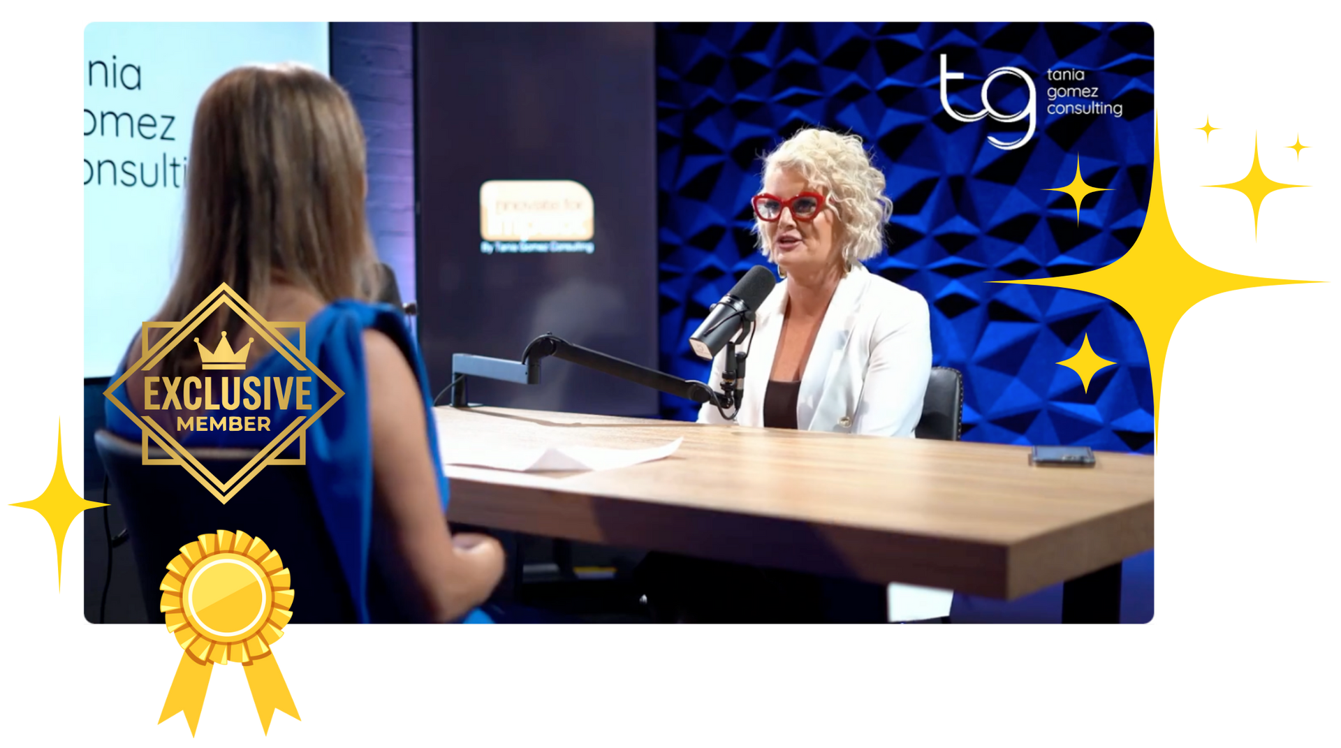 Interviewer at desk with woman in white jacket on blue stage background, TV interview setting