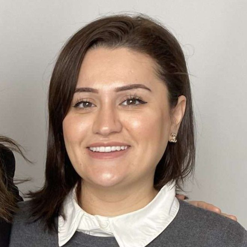 Smiling person with short brown hair, wearing a white-collared shirt and dark sweater against a plain background