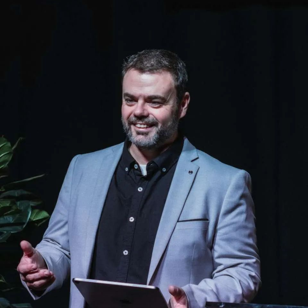 Man in a light gray blazer speaking on stage, holding notes and gesturing against a dark background