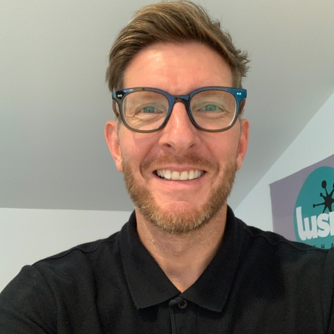 Smiling person wearing blue glasses and a black polo shirt in a bright indoor office.