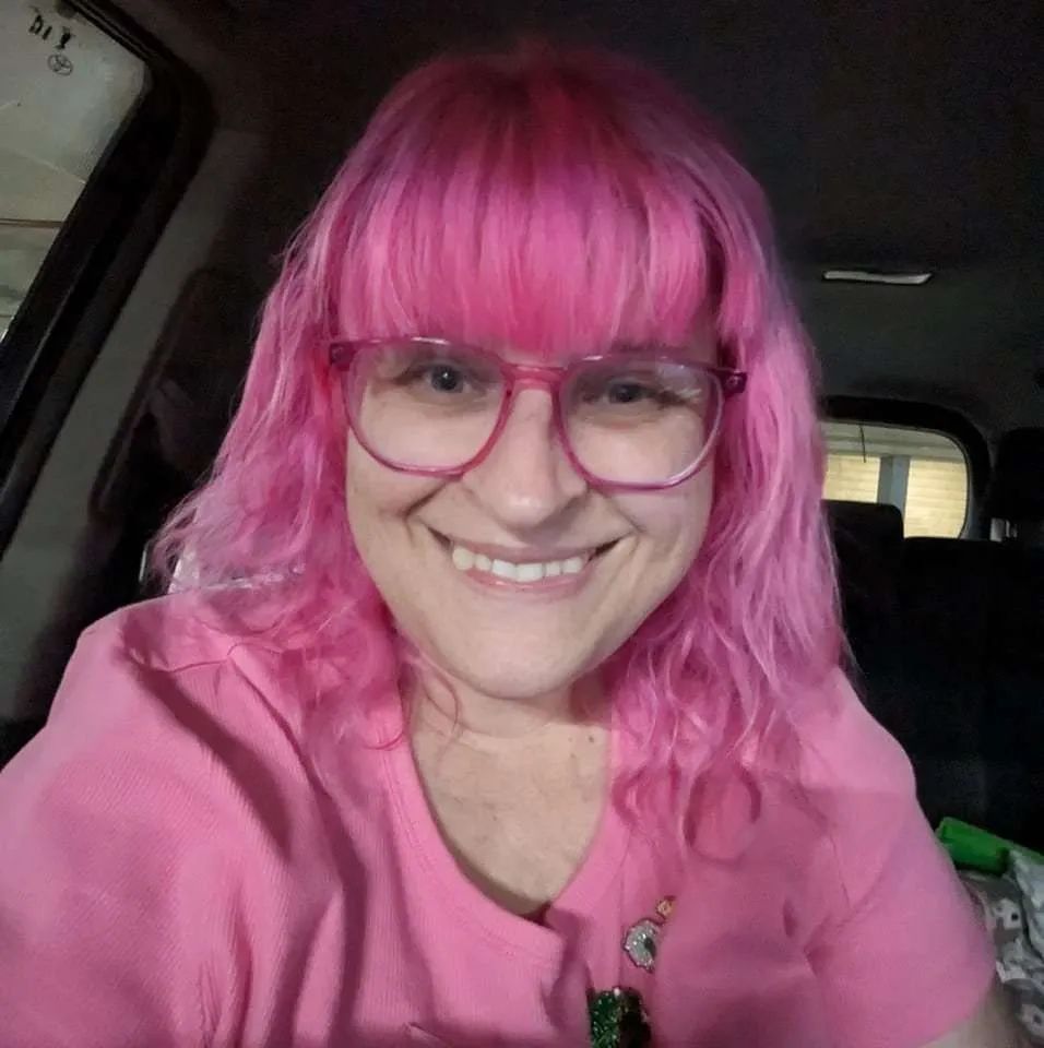 Smiling person with pink hair and glasses wearing a pink shirt inside a car