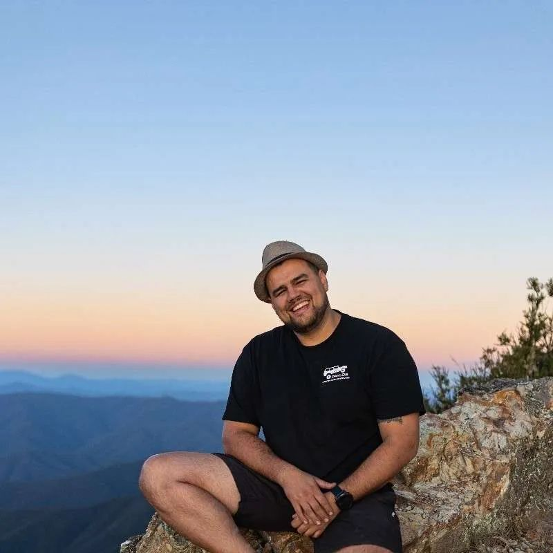 Smiling person in a hat sitting on a rocky overlook at sunset with mountains in the background