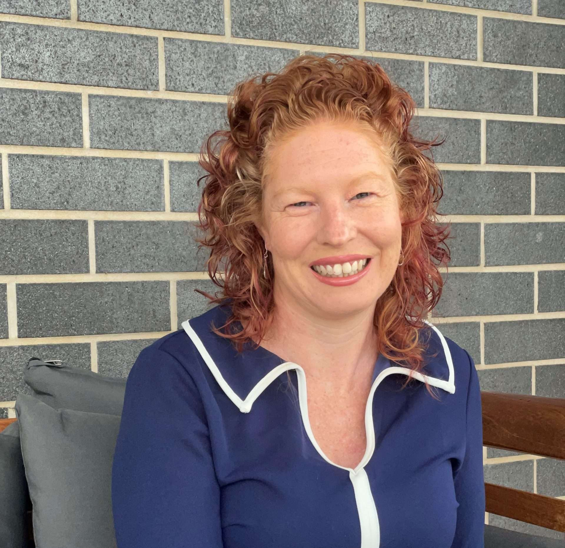 Smiling woman with curly red hair in a navy top sitting against a gray brick wall
