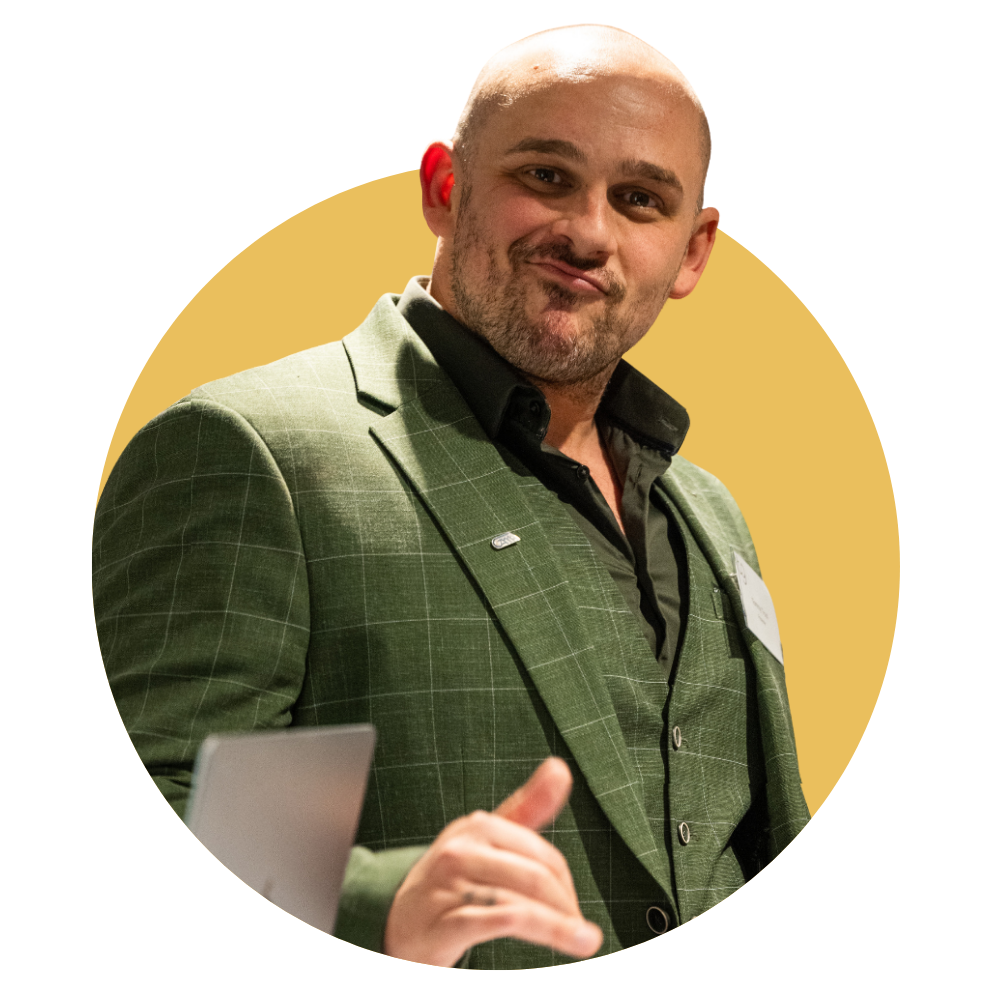 Man in a green blazer smiling and gesturing in front of a yellow circular backdrop