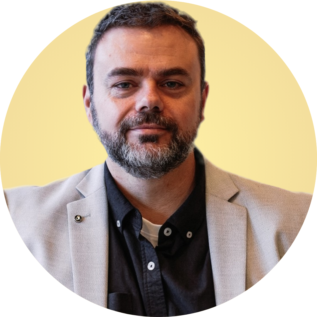 Portrait of a bearded man in a light blazer and dark shirt against a pale yellow background