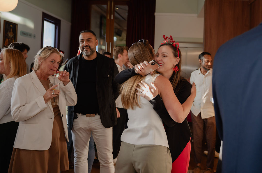 People hugging and chatting at an indoor gathering, with drinks and a small audience in the background.