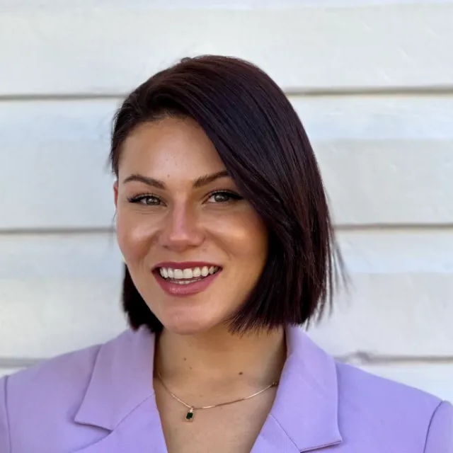 Smiling person with short dark hair in a lavender blazer against a white wall