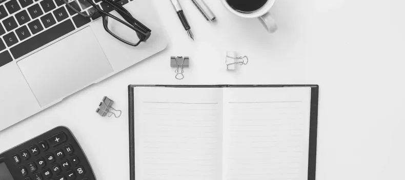 Top-down desk workspace with open notebook, laptop, calculator, and paper clips on a white surface