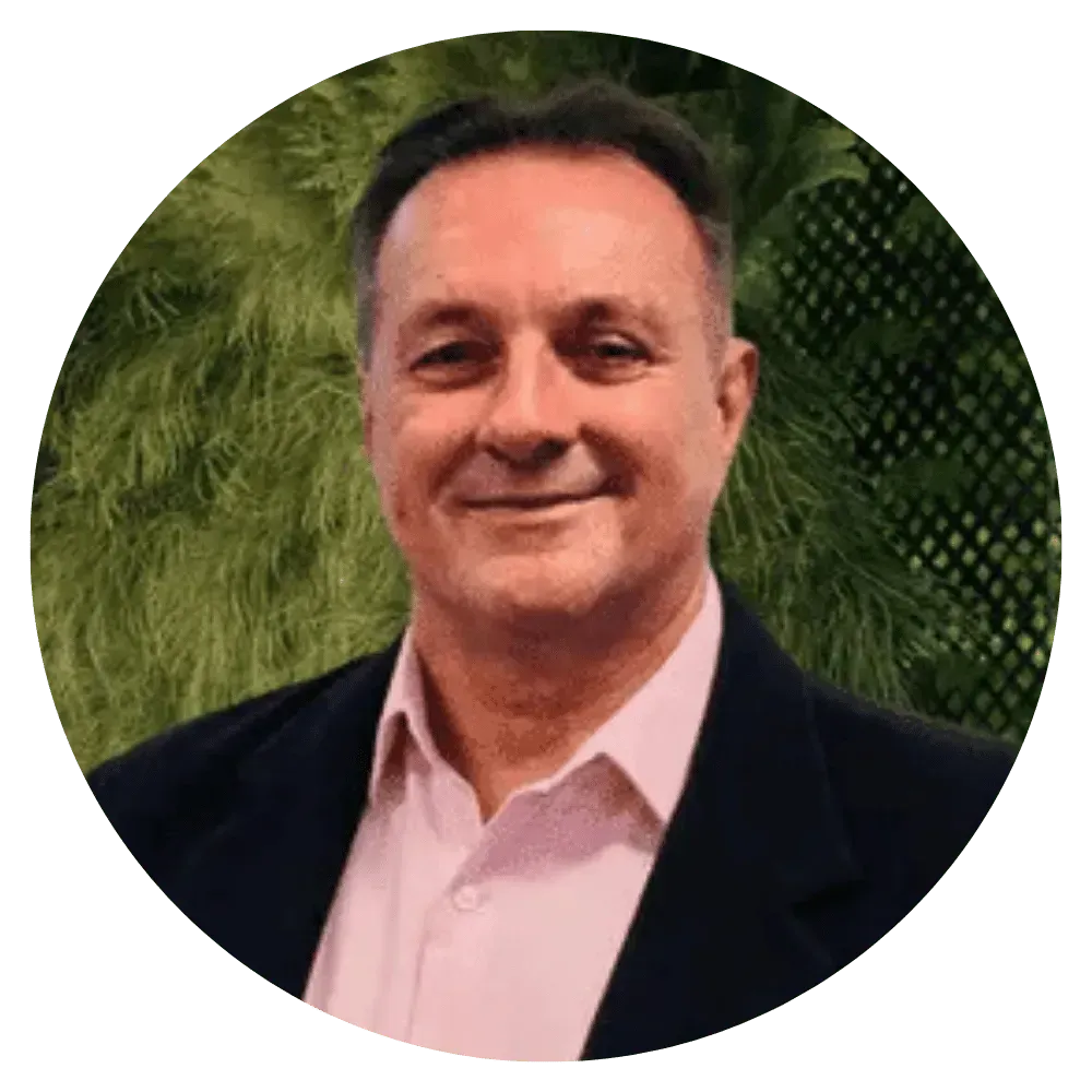Portrait of a smiling man in a dark blazer and pink shirt against a green leafy background