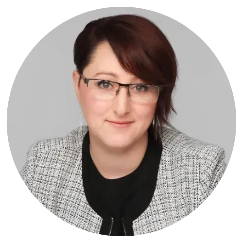 Headshot of a person with short dark hair and glasses, wearing a gray blazer over a black top.