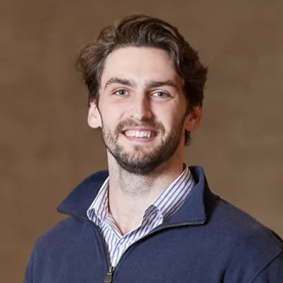 Smiling man in a navy sweater over a striped shirt against a plain brown background