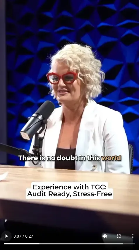 Woman speaking at a podium with microphone on a blue-lit stage, wearing red glasses and a white blazer.
