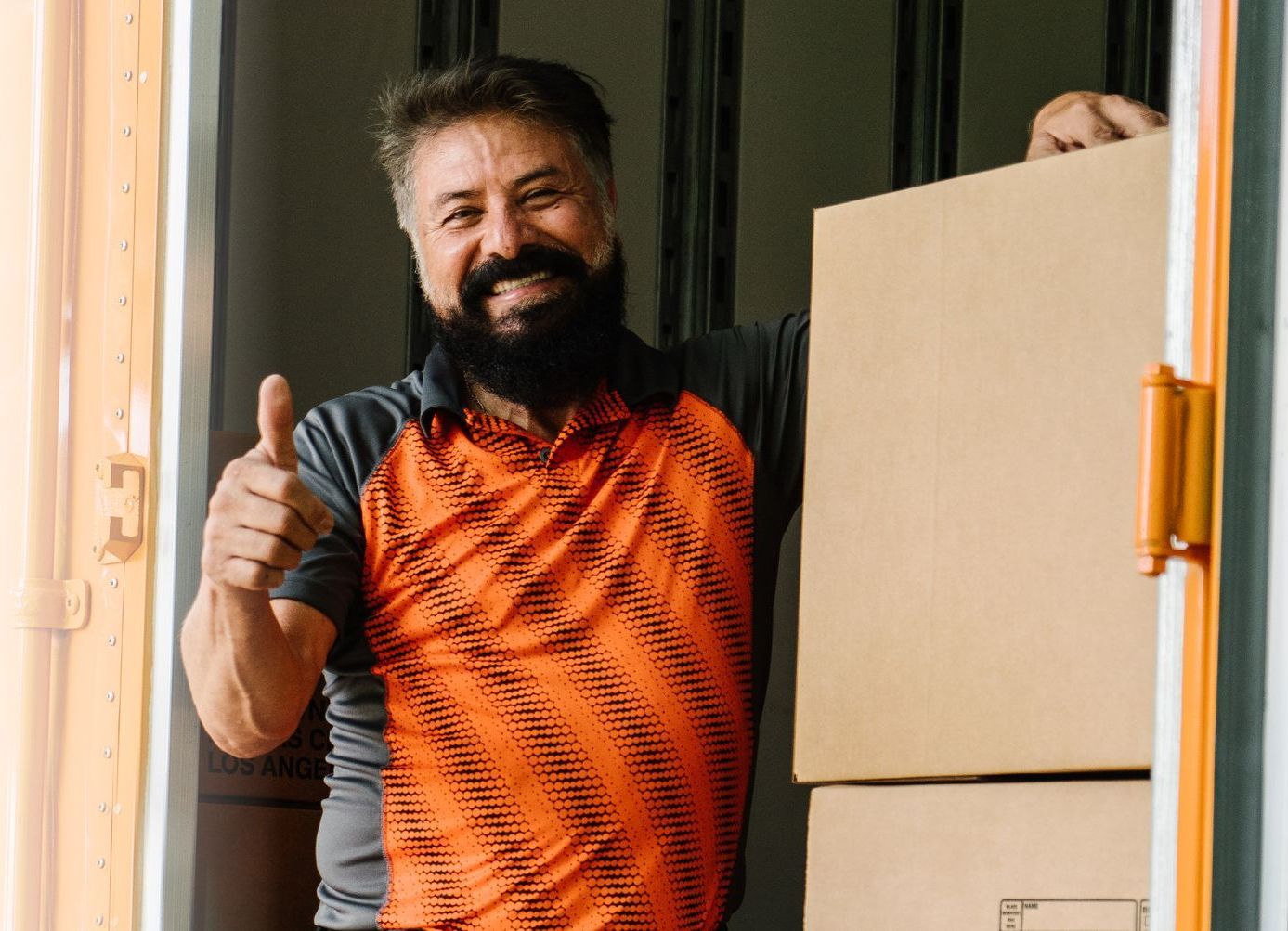 Un homme barbu, le pouce levé, tient des cartons à l'intérieur d'un camion de déménagement et sourit.