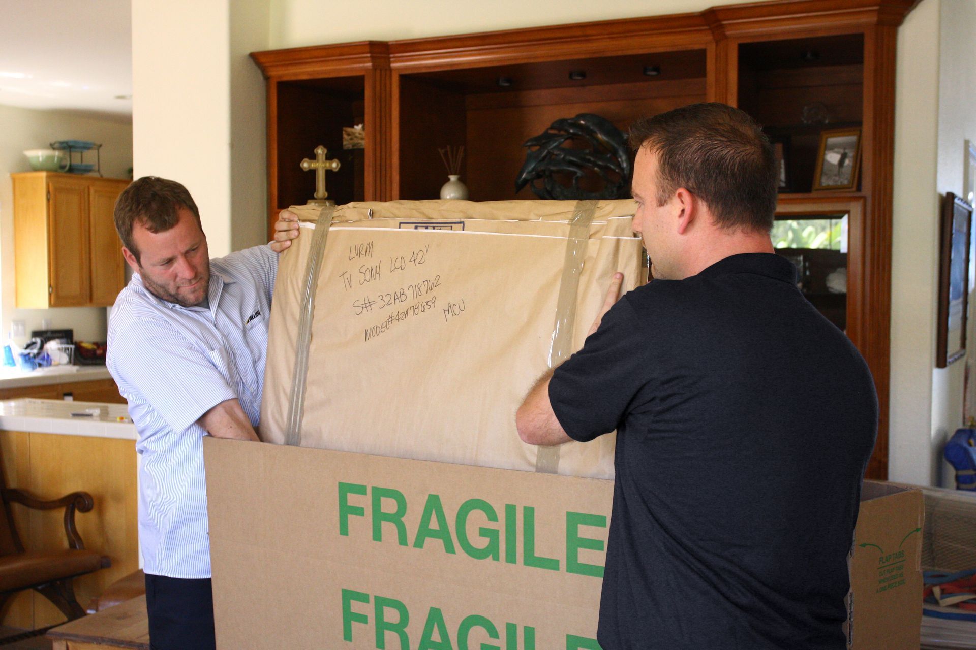Deux personnes soulèvent un grand carton portant l'inscription « FRAGILE » à l'intérieur d'une maison. Tons bruns et beiges.