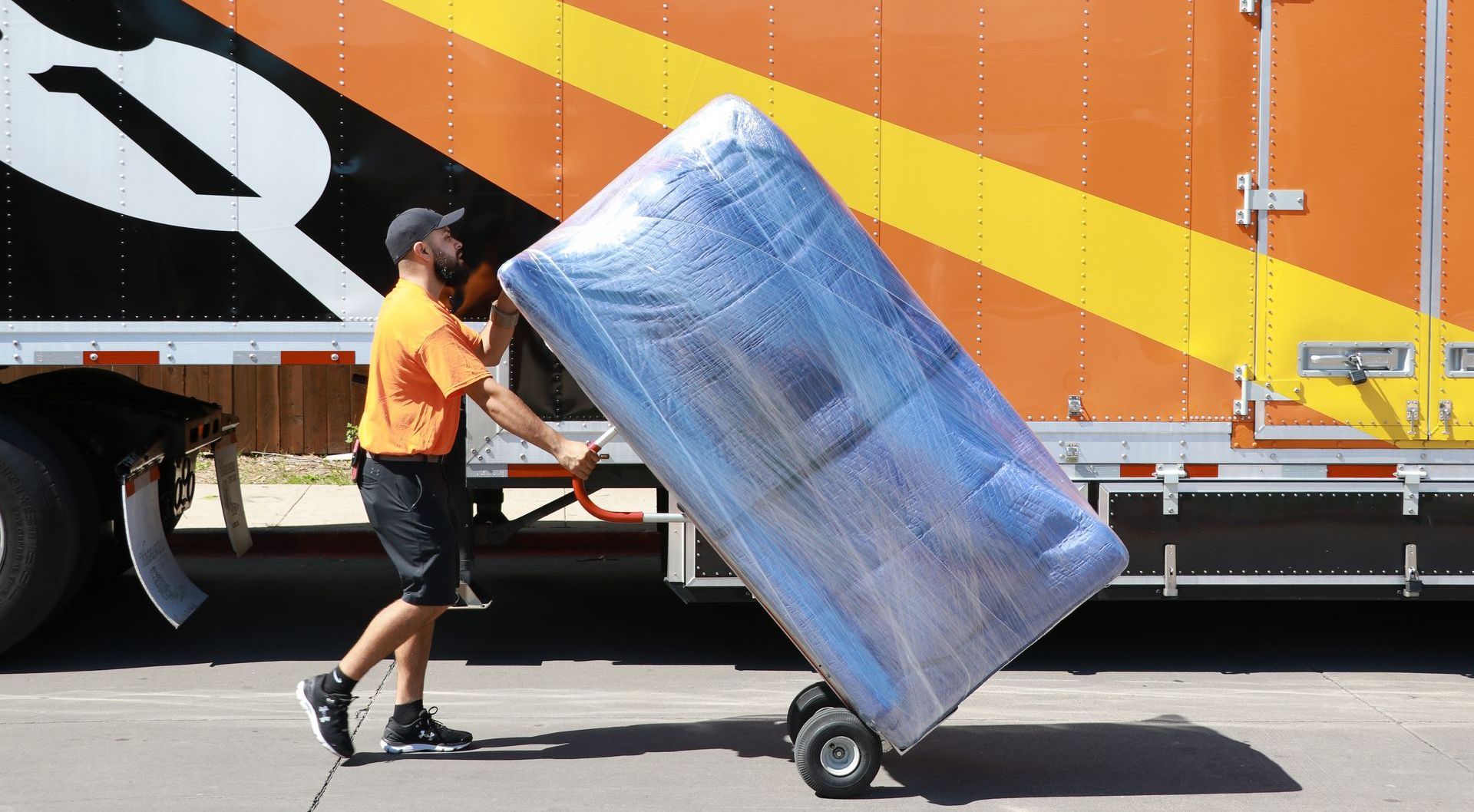 Un homme en chemise orange pousse un matelas emballé sur un chariot vers un grand camion de déménagement orange.