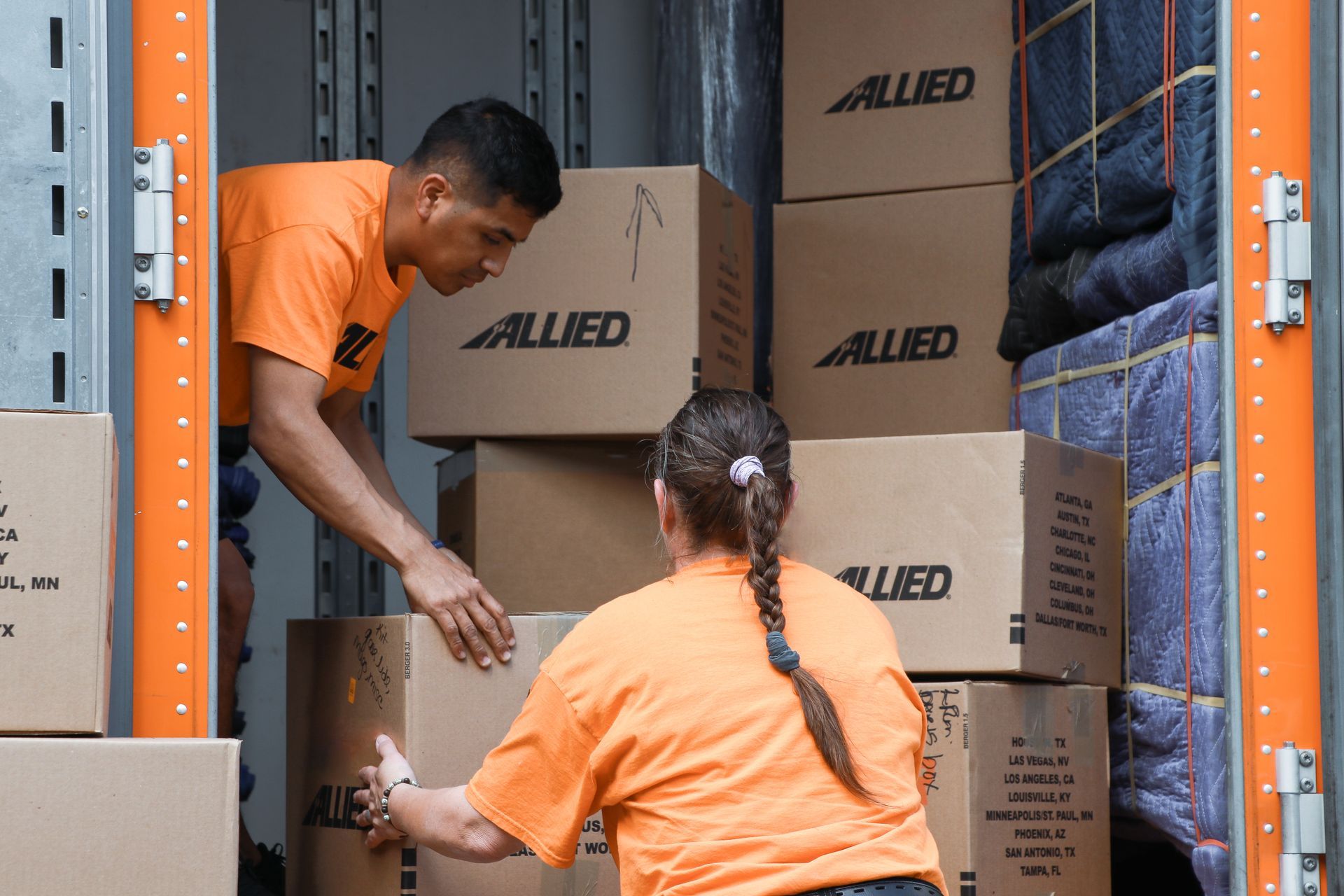 Deux personnes en chemises orange chargent des cartons dans un camion de déménagement, cartons étiquetés « ALLIÉS ».