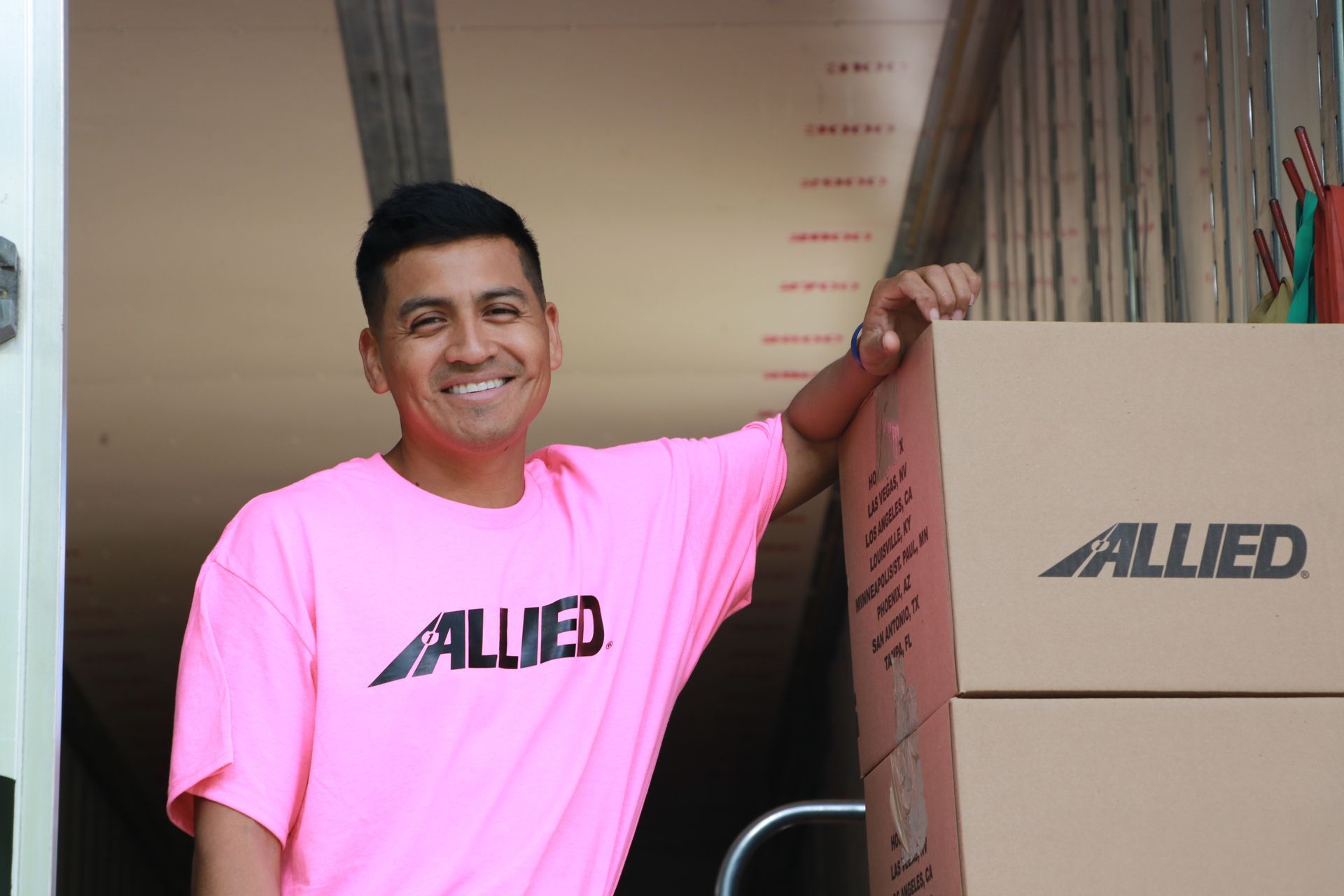 Un homme portant un t-shirt rose « Alliés », souriant, s'appuie sur des cartons de déménagement à l'intérieur d'un camion.