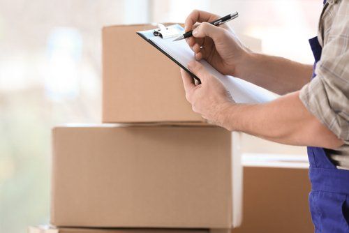 A man is writing on a clipboard in front of a stack of cardboard boxes.