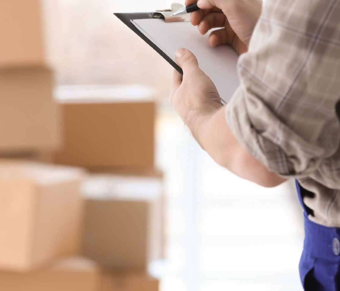 A man is writing on a clipboard with boxes in the background