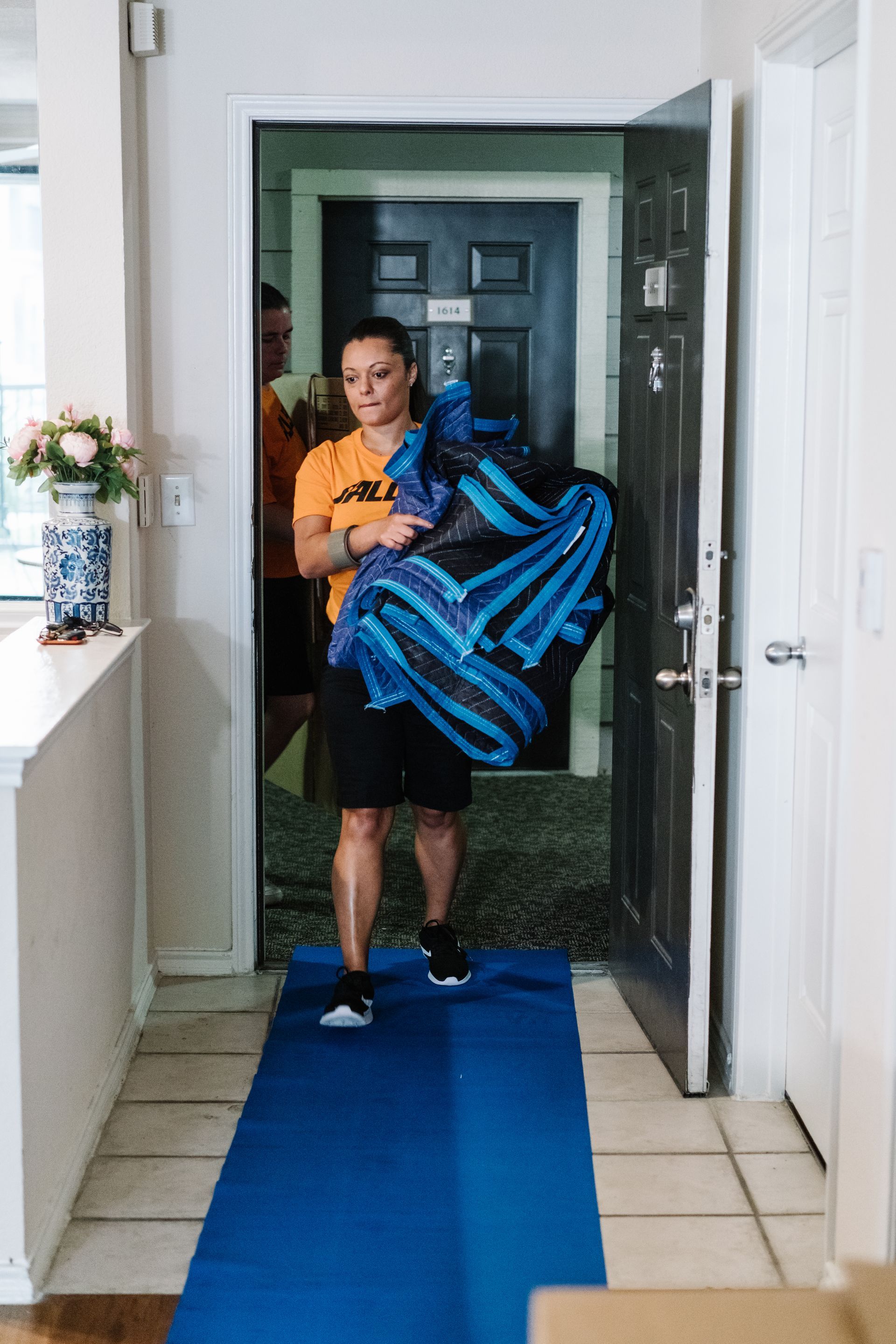 Une femme portant un grand sac bleu et noir se tient debout dans l'embrasure d'une porte. Elle est assise sur un tapis bleu à l'intérieur de l'embrasure.