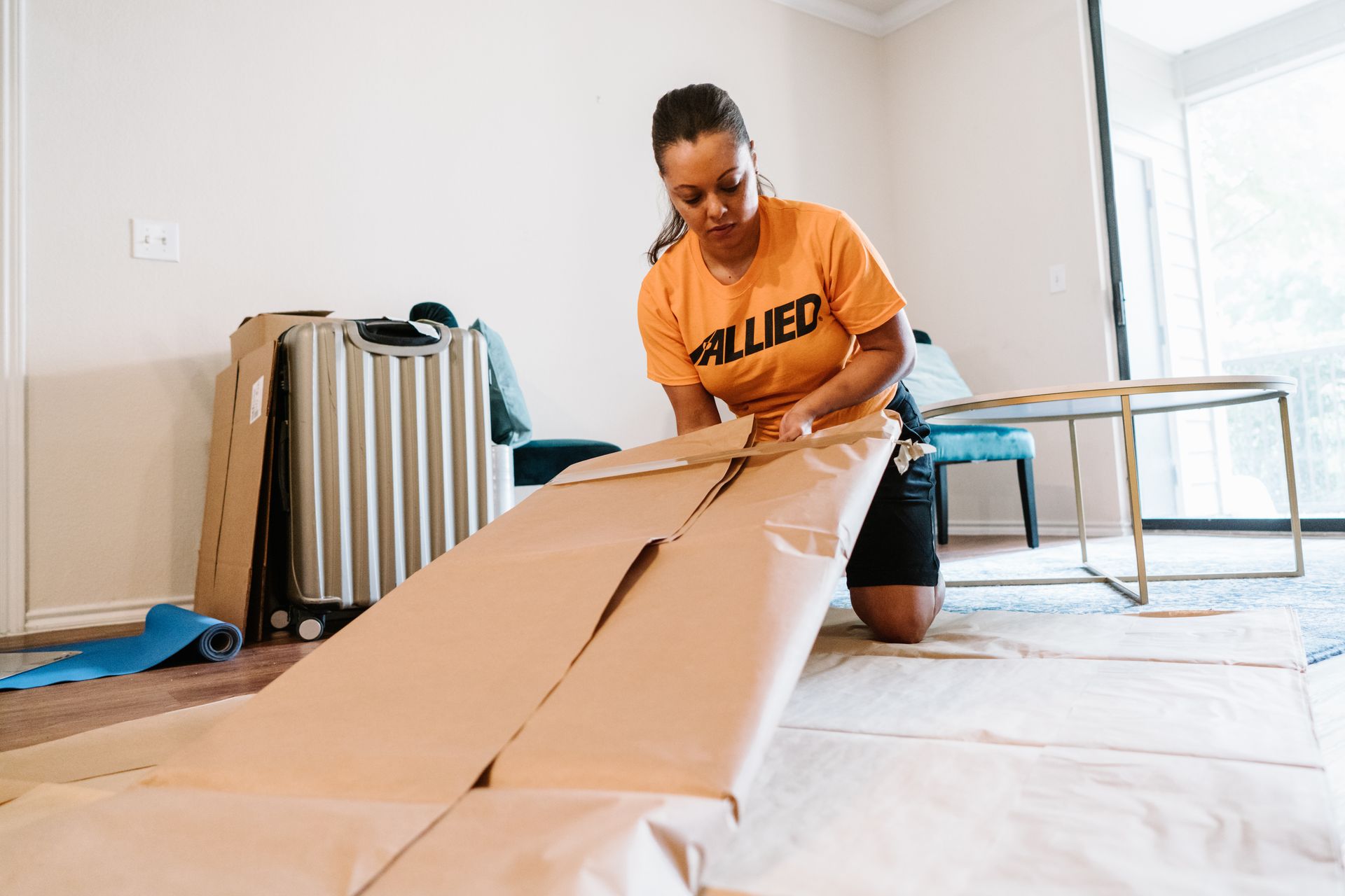 Une femme est agenouillée sur le sol, en train d'emballer un objet dans du papier kraft, dans une pièce où se trouvent des bagages et des meubles.